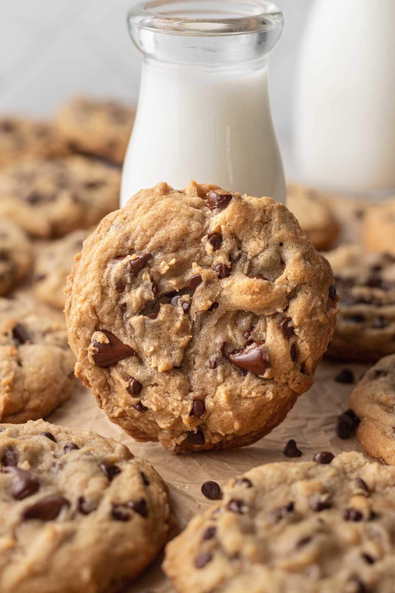 Una gran galleta con chispas de chocolate estilo panadería apoyada contra un vaso alto de leche.  Más galletas lo rodean.