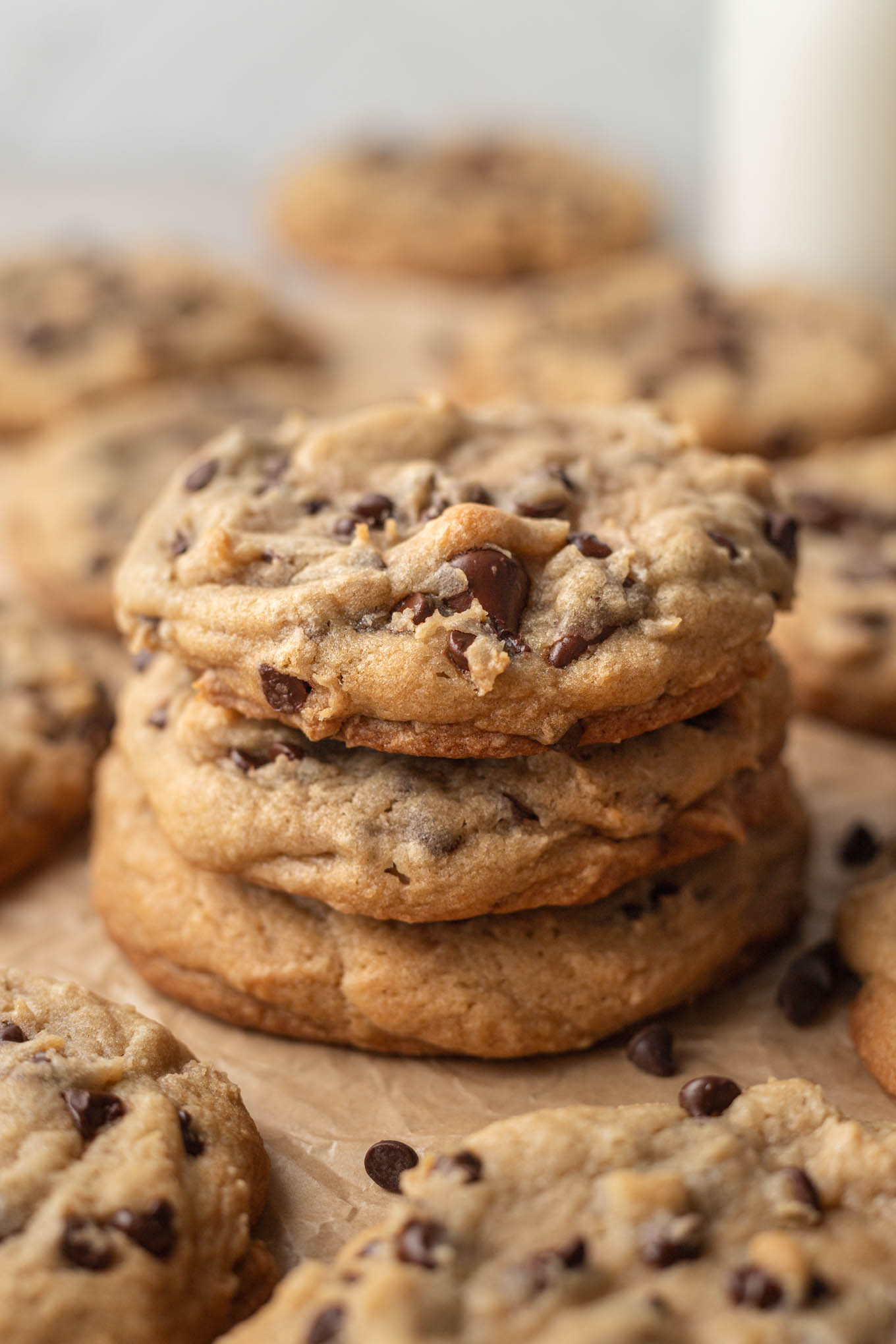 Una pila de tres galletas con chispas de chocolate estilo panadería sobre un trozo de papel pergamino marrón.  Hay más cookies rodeando la pila de cookies.