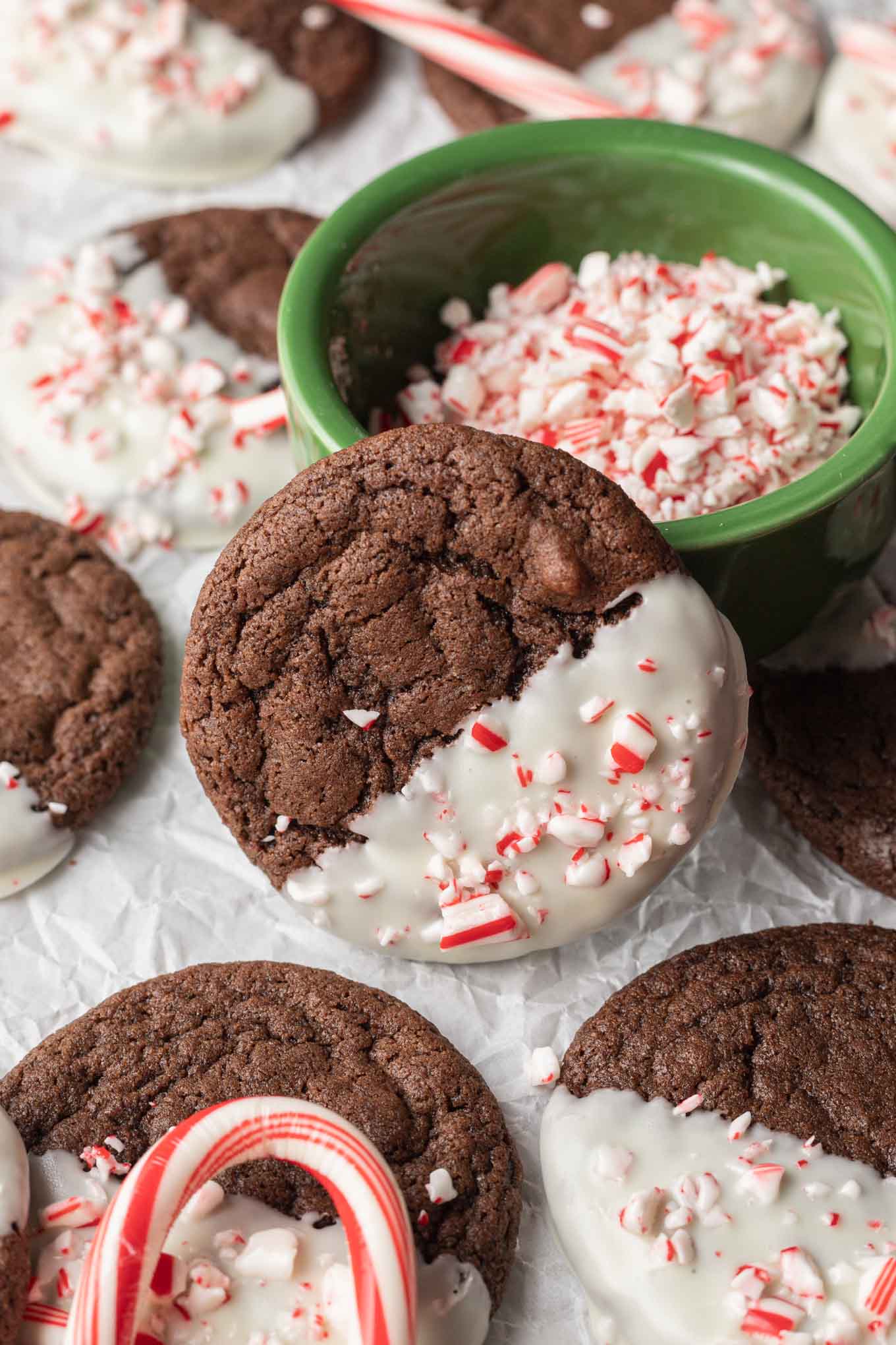 Una galleta de chocolate con menta apoyada sobre un tazón con trozos de bastón de caramelo, rodeada de galletas adicionales. 