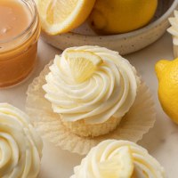 Cupcakes de limón cubiertos con crema de mantequilla de limón y una rodajita de limón. Un tazón de limones y un frasco de crema de limón se encuentran junto a los pastelitos.
