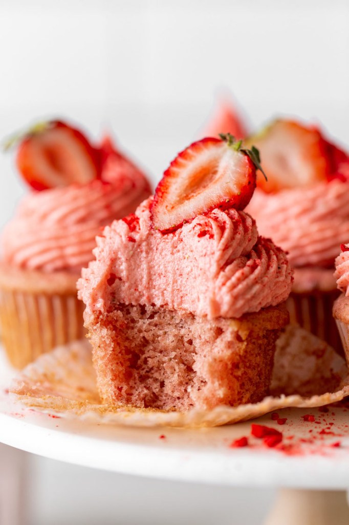 Una vista de cerca de pastelitos con glaseado de fresa en un puesto de pasteles.  Al cupcake del frente le falta un bocado. 