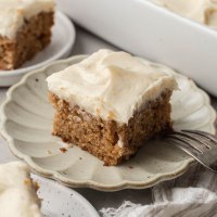 Una rebanada de pastel de puré de manzana cubierta con glaseado de queso crema y mantequilla marrón en un plato festoneado.