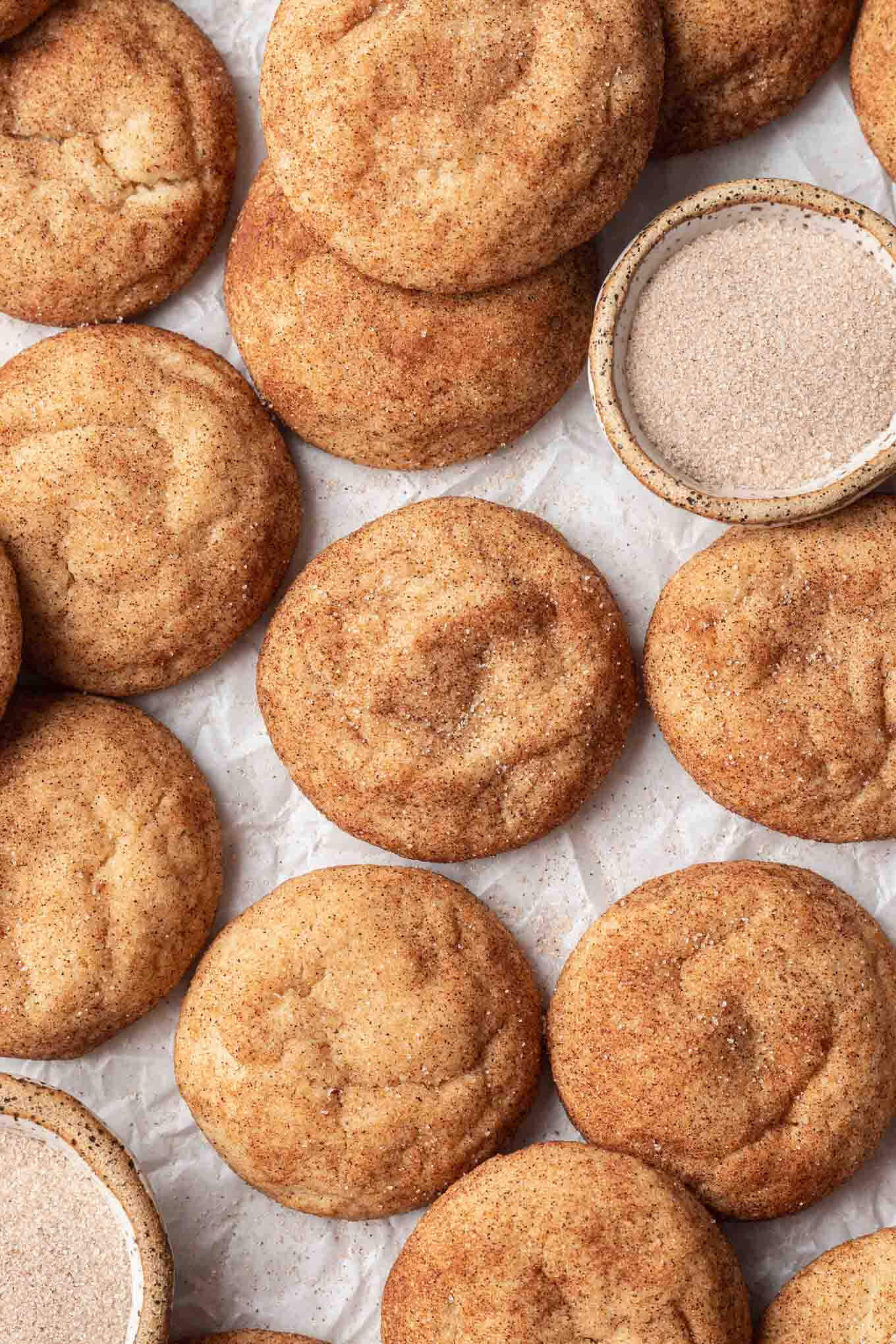 Una vista aérea de snickerdoodles de mantequilla dorada sobre papel pergamino, con un plato de azúcar y canela a un lado. 