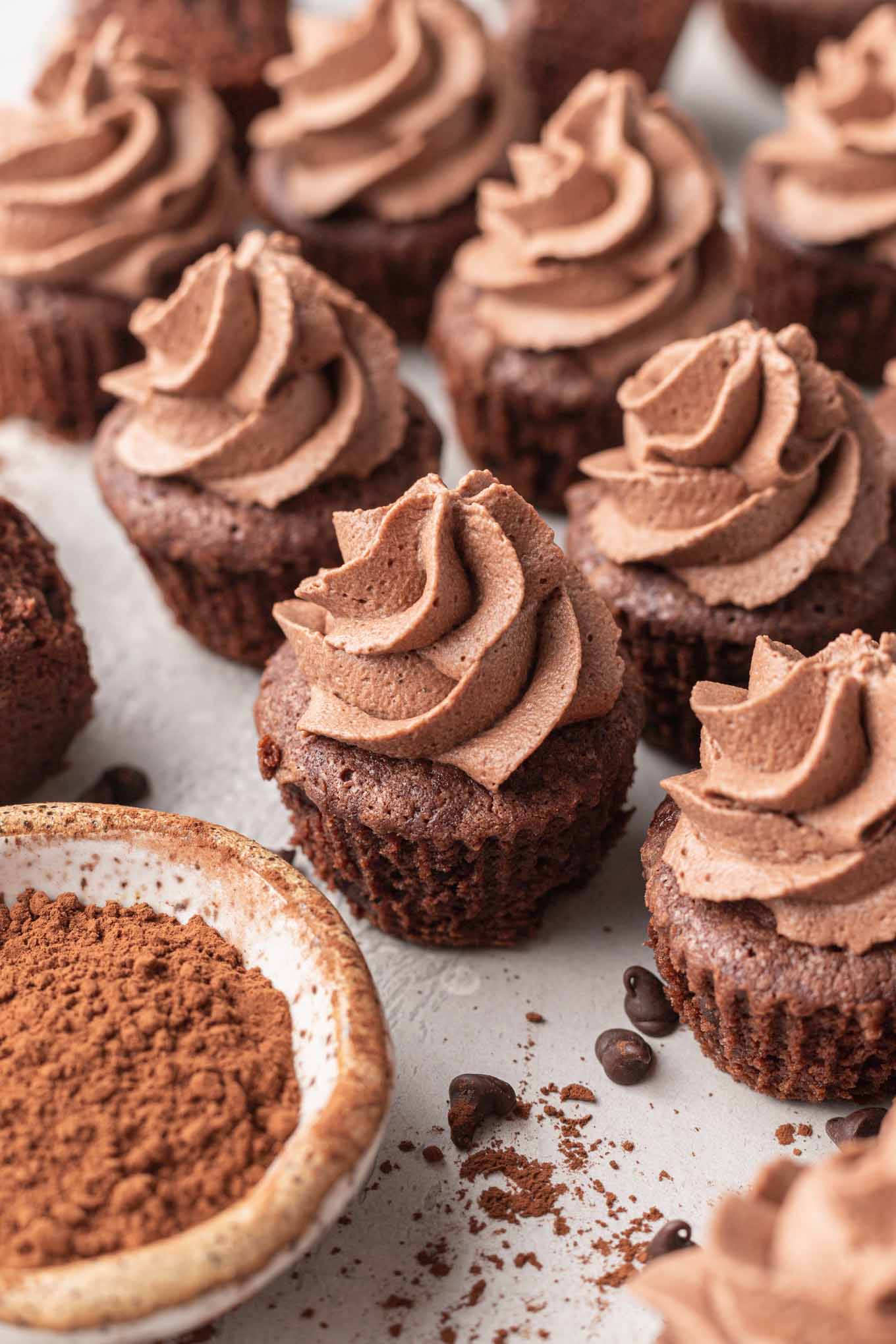 Mini brownies cubiertos con crema batida de chocolate, junto a un bol de cacao en polvo. 