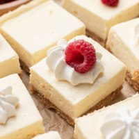 Varias barras de tarta de queso en rodajas sobre un trozo de papel pergamino.  Algunas de las barras están cubiertas con crema batida y frambuesa.