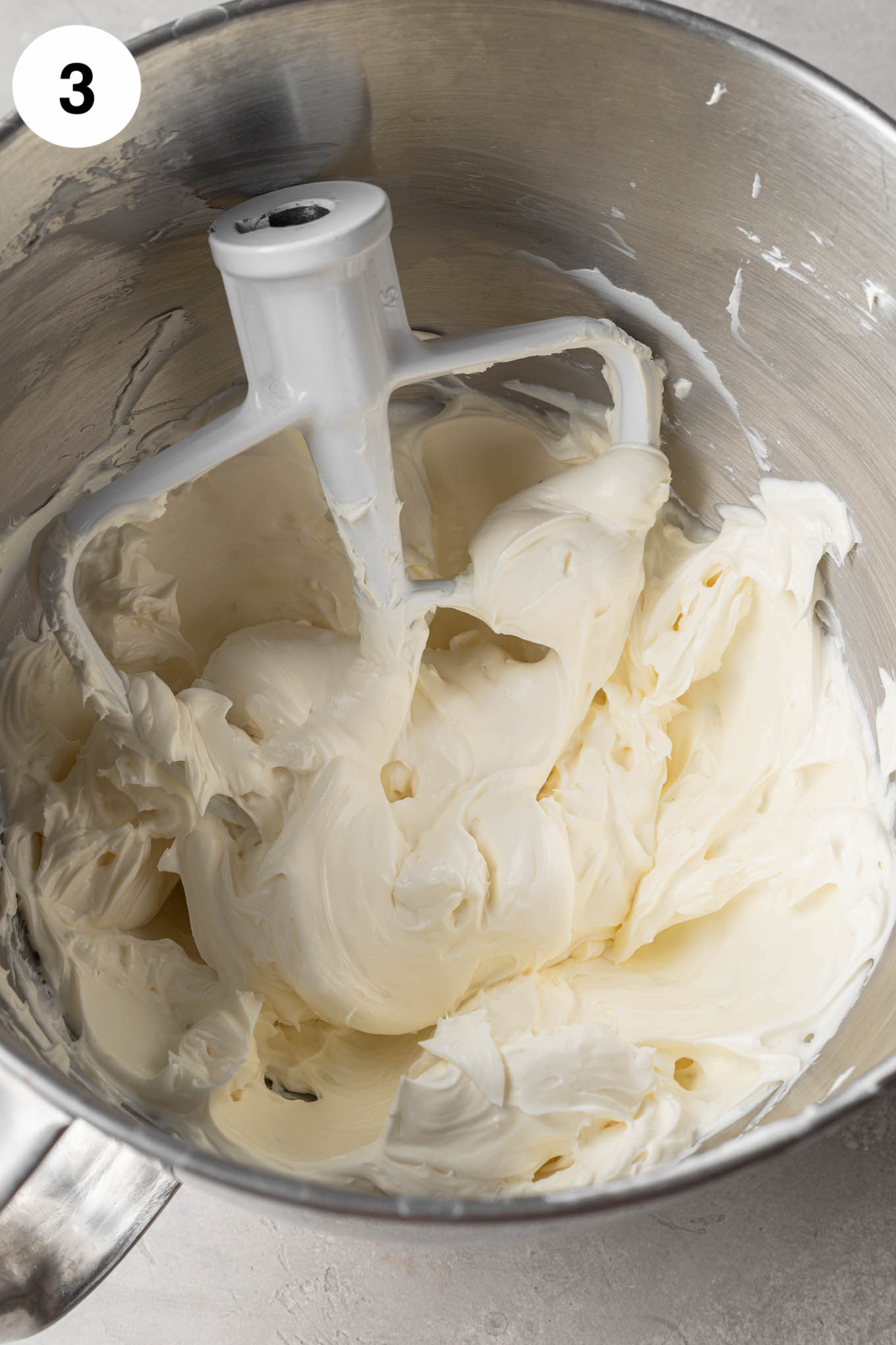 Una vista aérea de queso crema batido, crema agria, azúcar y vainilla en el tazón de una batidora de pie, con un accesorio de paleta. 