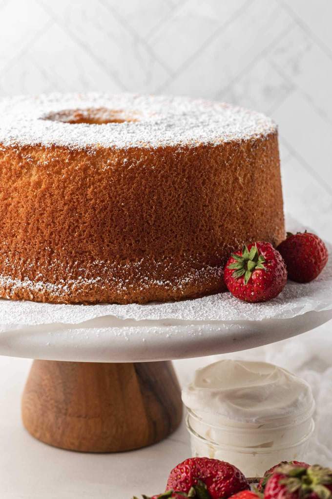 Un pastel de gasa horneado encima de un soporte para pasteles de mármol.  El pastel ha sido espolvoreado con azúcar en polvo y al lado hay fresas frescas.