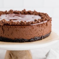 A chocolate cheesecake sitting on top of a marble cake stand. The cheesecake is topped with chocolate ganache and garnished with chocolate curls.