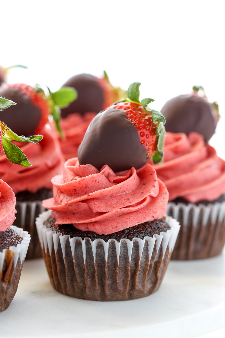 Un primer plano de pastelitos de chocolate cubiertos con glaseado de crema de mantequilla de fresa y fresas cubiertas de chocolate en un soporte de pastel de mármol blanco.