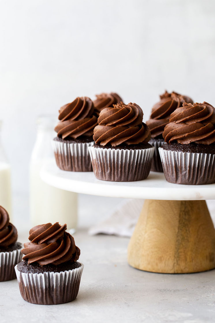 Un puesto de pasteles de mármol que sostiene pastelitos de chocolate cubiertos con glaseado de chocolate y jarras de leche al fondo.