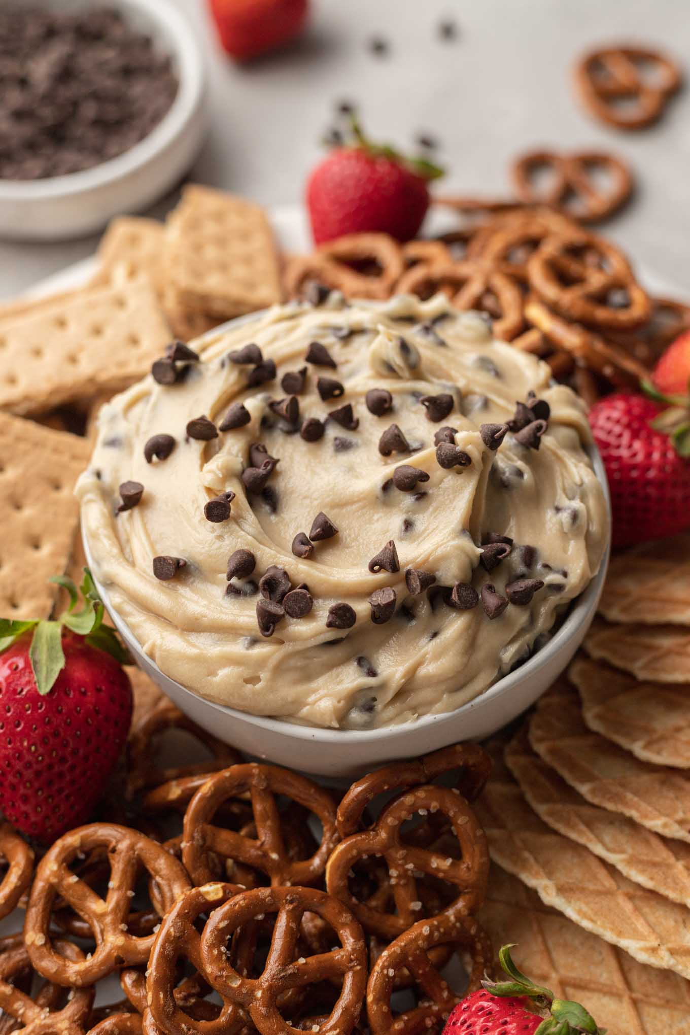 Un plato de salsa de masa para galletas de queso crema, rodeado de trozos de cono de waffle, pretzels, bayas y galletas integrales. 