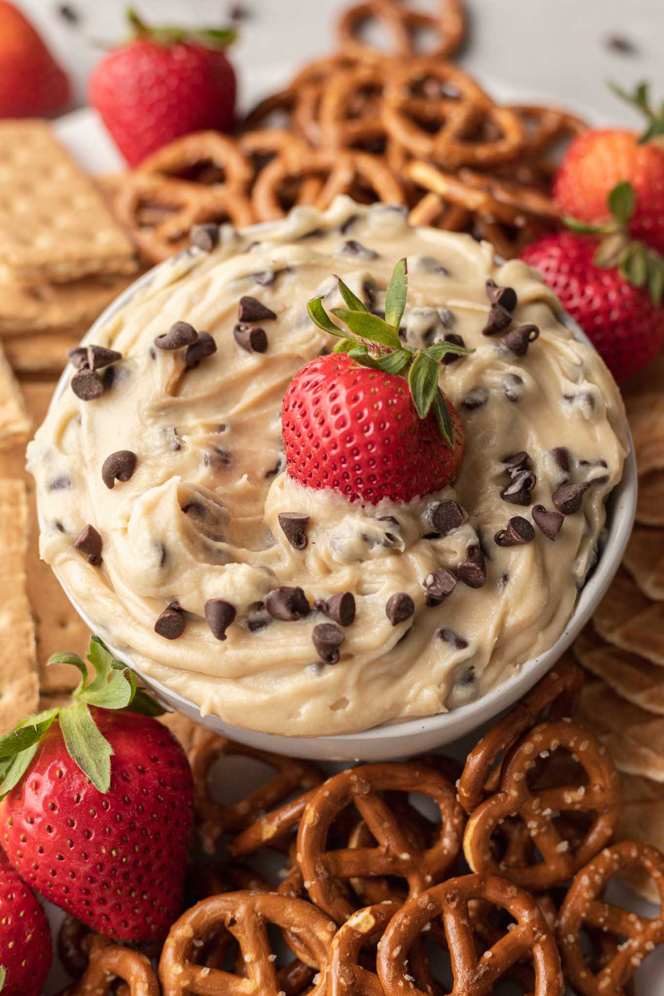 Un plato de salsa de masa para galletas, rodeado de trozos de cono de waffle, pretzels, bayas y galletas integrales. 