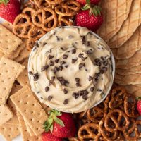 Una vista aérea de la masa para galletas en un tazón.  El cuenco está rodeado de pretzels, galletas integrales, fresas y trozos de conos de waffle.