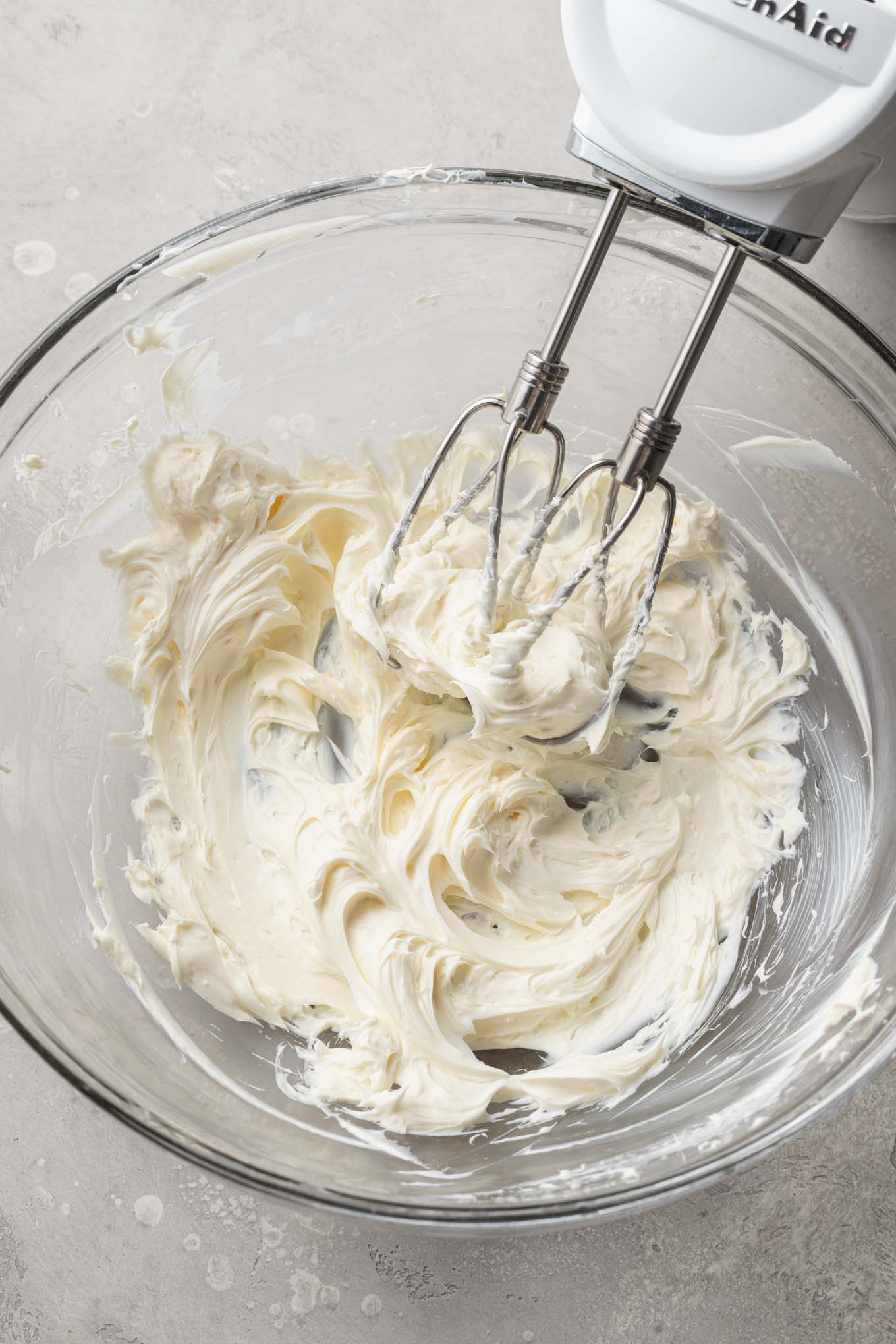 Una vista aérea del queso crema batido en un tazón de vidrio para mezclar, con una batidora de mano. 