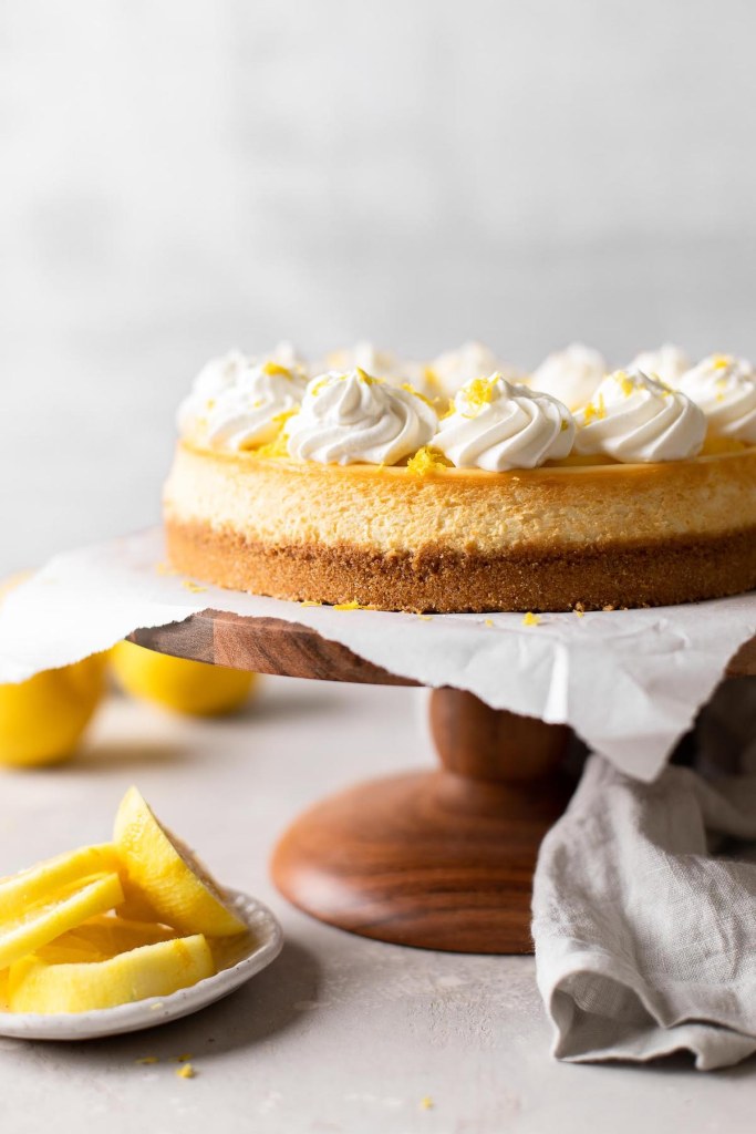 Una vista lateral de una tarta de queso con limón sobre un soporte para tartas de madera.  Las rodajas de limón descansan en un plato pequeño cercano. 