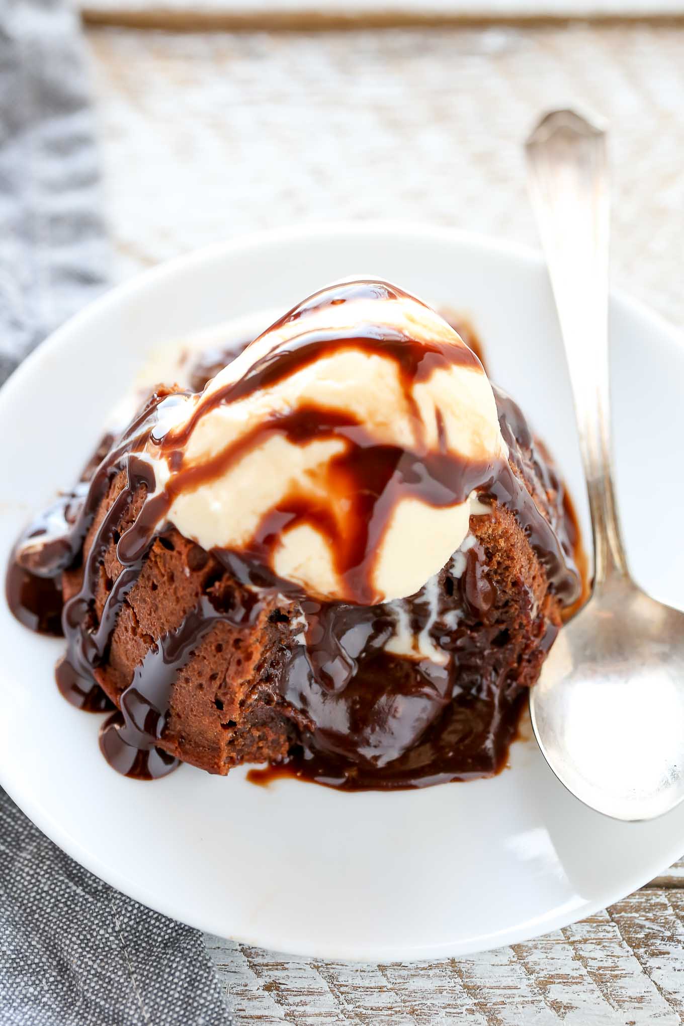 Un pastel de lava fundida cubierto con helado y salsa de chocolate en un plato blanco.  Se ha cortado el bizcocho y el relleno rezuma. 