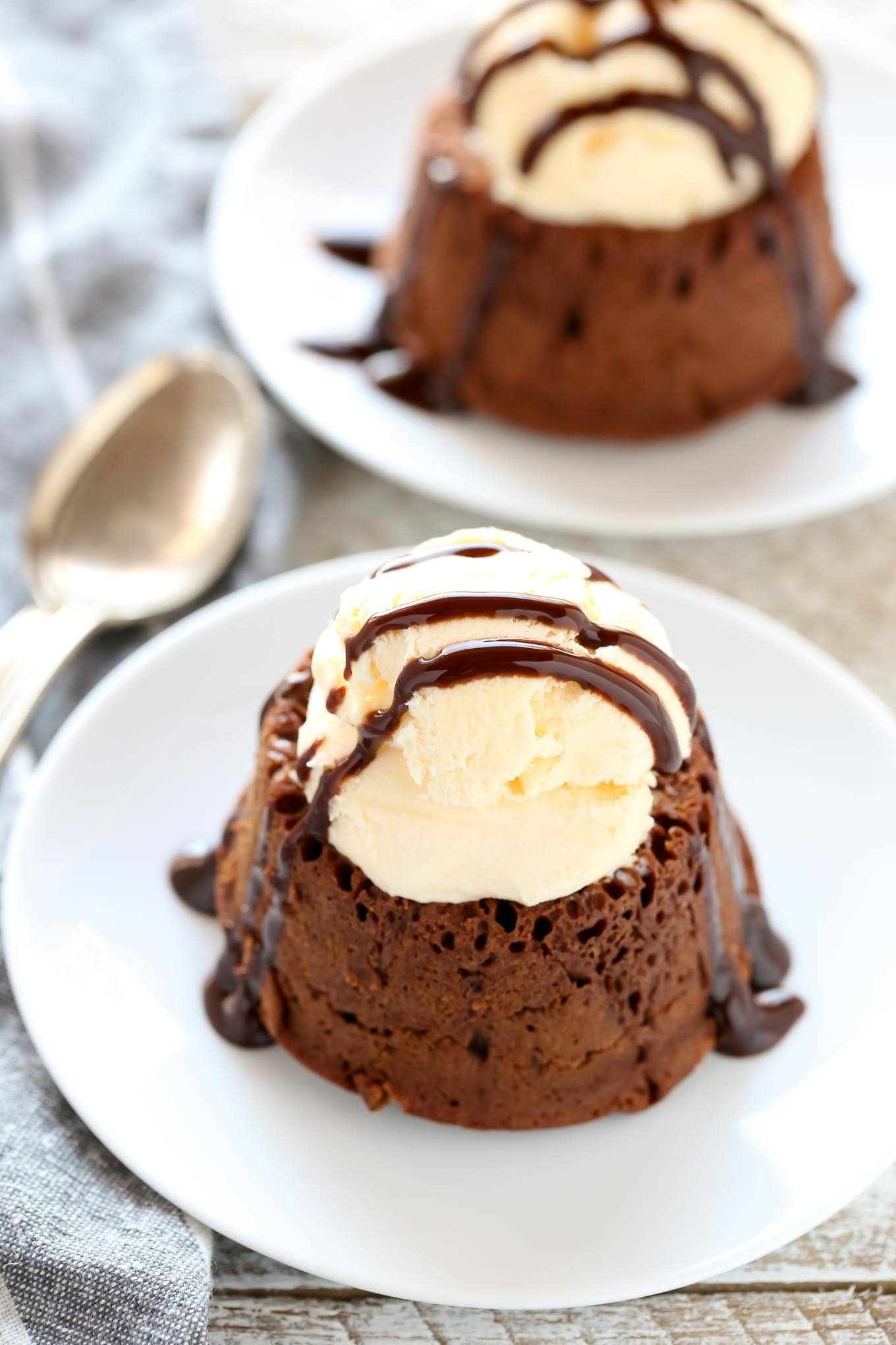 Dos lava cakes de chocolate cubiertos con una bola de helado y un chorrito de salsa de chocolate.  Ambos están en platos de postre blancos. 