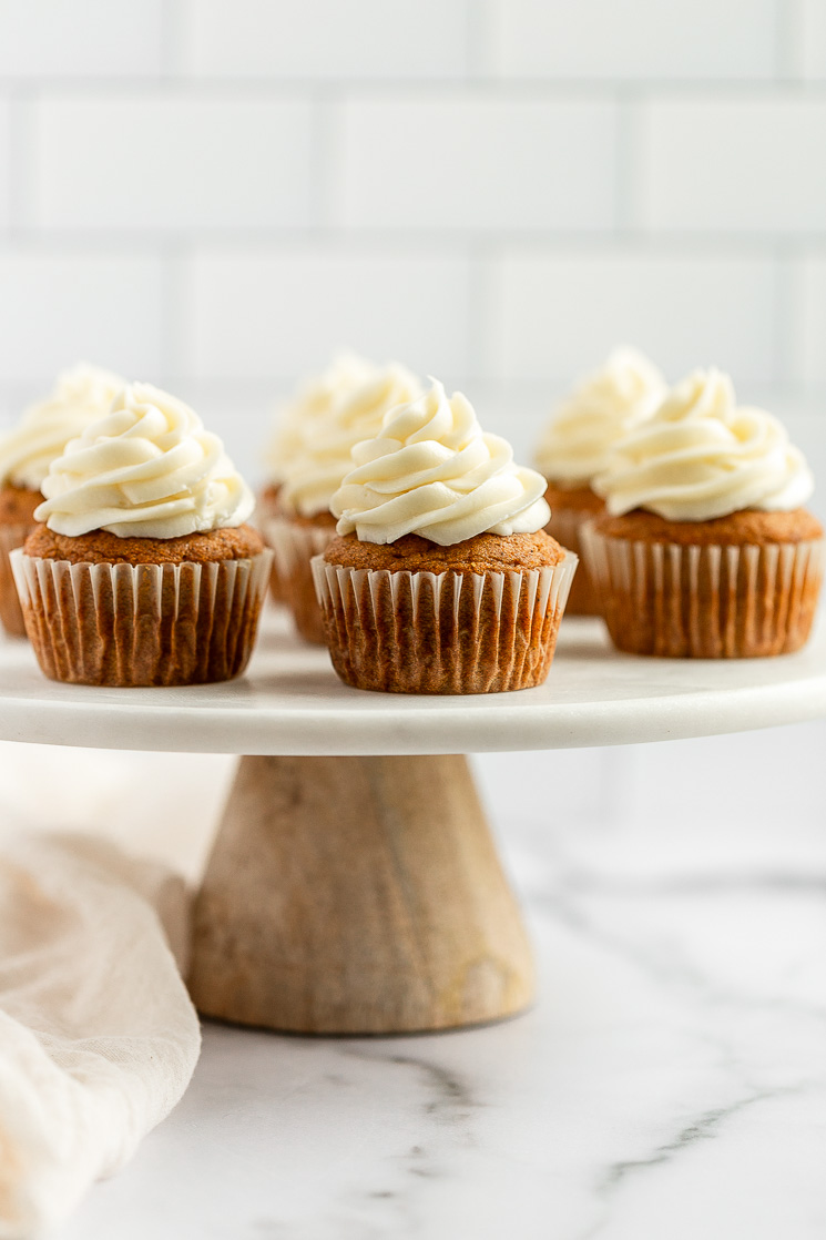 Un grupo de pastelitos de calabaza cubiertos con glaseado de queso crema colocados encima de un soporte para pasteles de mármol.