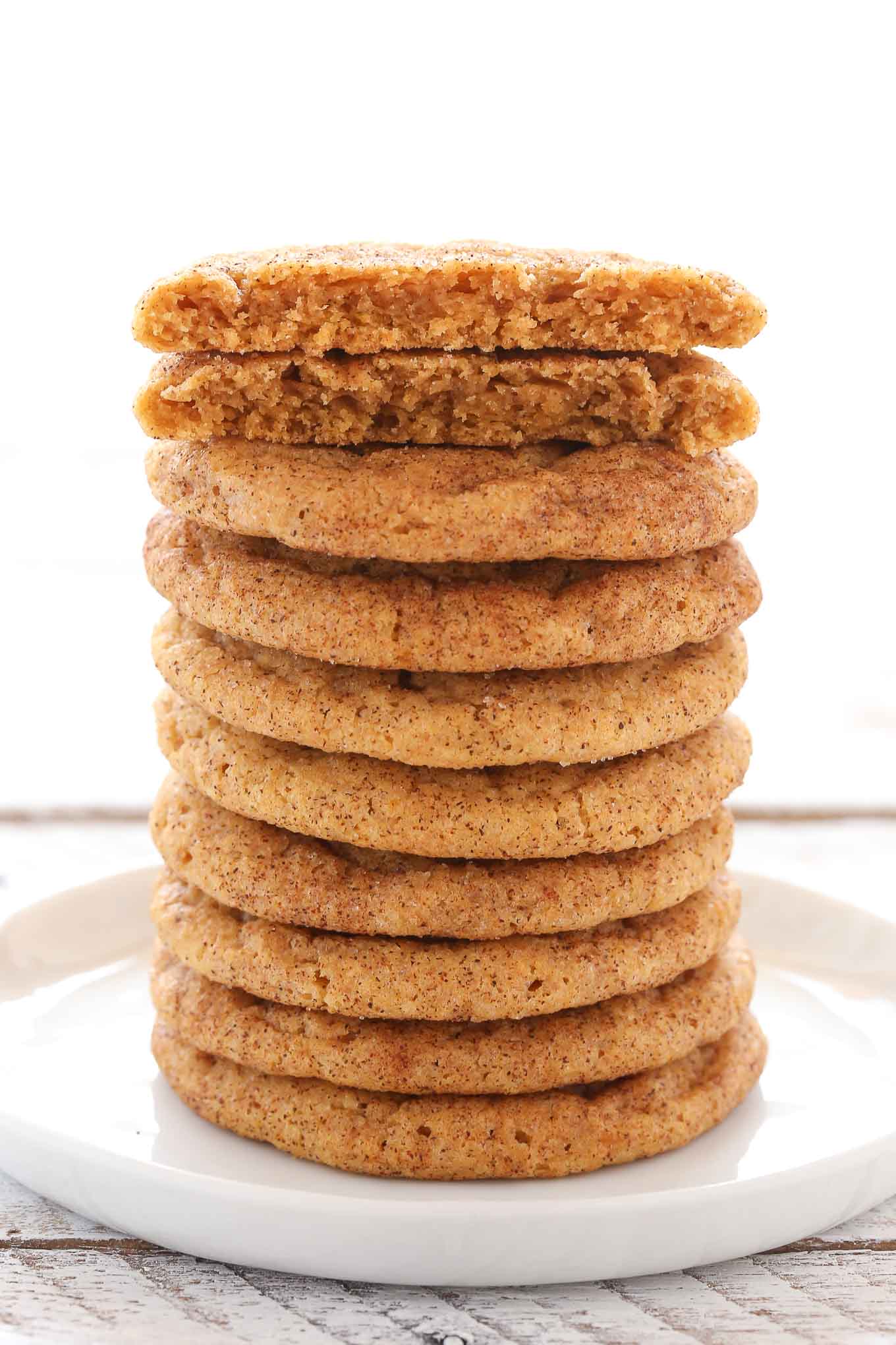 Una pila alta de snickerdoodles de calabaza en un plato blanco. La galleta superior se partió por la mitad.