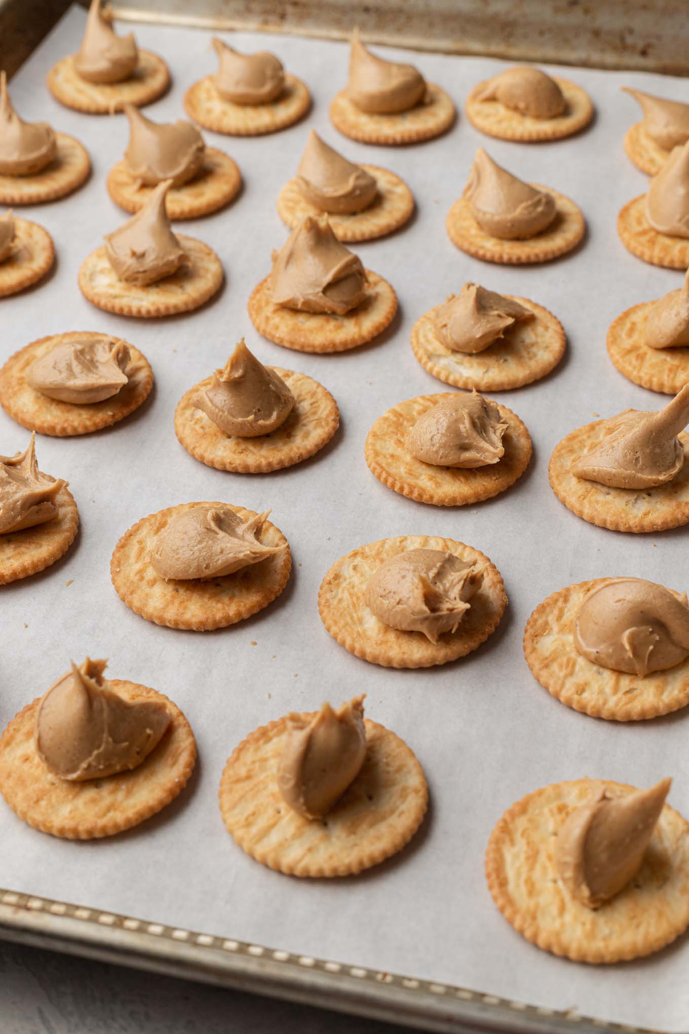 Filas de galletas saladas cubiertas con relleno de mantequilla de maní, alineadas en una bandeja para hornear. 