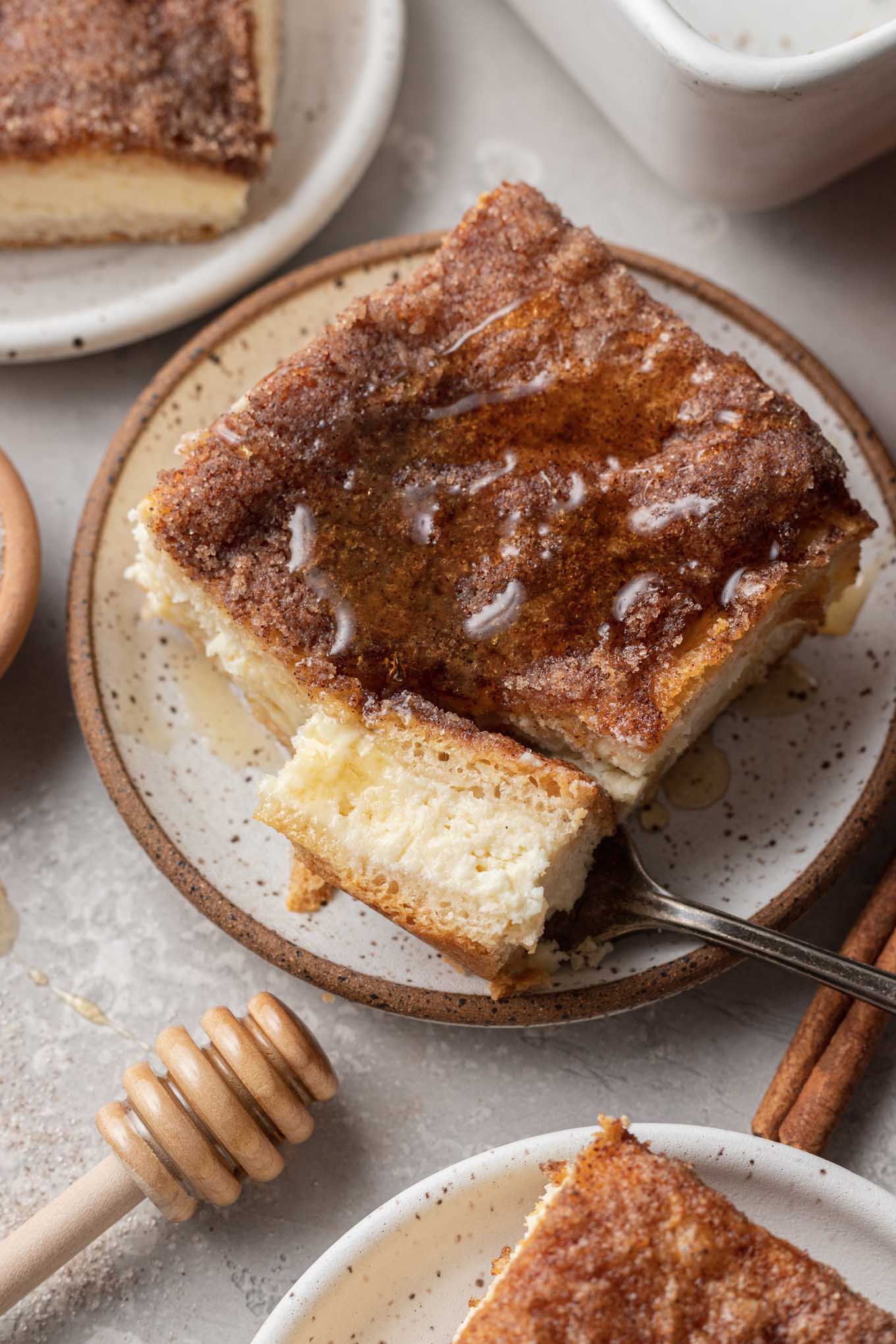 Una rebanada de cheesecake de sopapilla, rociada con miel.  Se ha pinchado un trozo con un tenedor que reposa sobre el plato. 