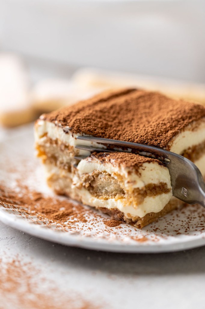 Una rodaja de tiramisú sobre un plato decorativo blanco.  Un tenedor se clava en el extremo de la rebanada.