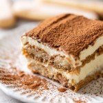 Una rodaja de tiramisú sobre un plato decorativo blanco.  Tres dedos femeninos descansan al fondo.