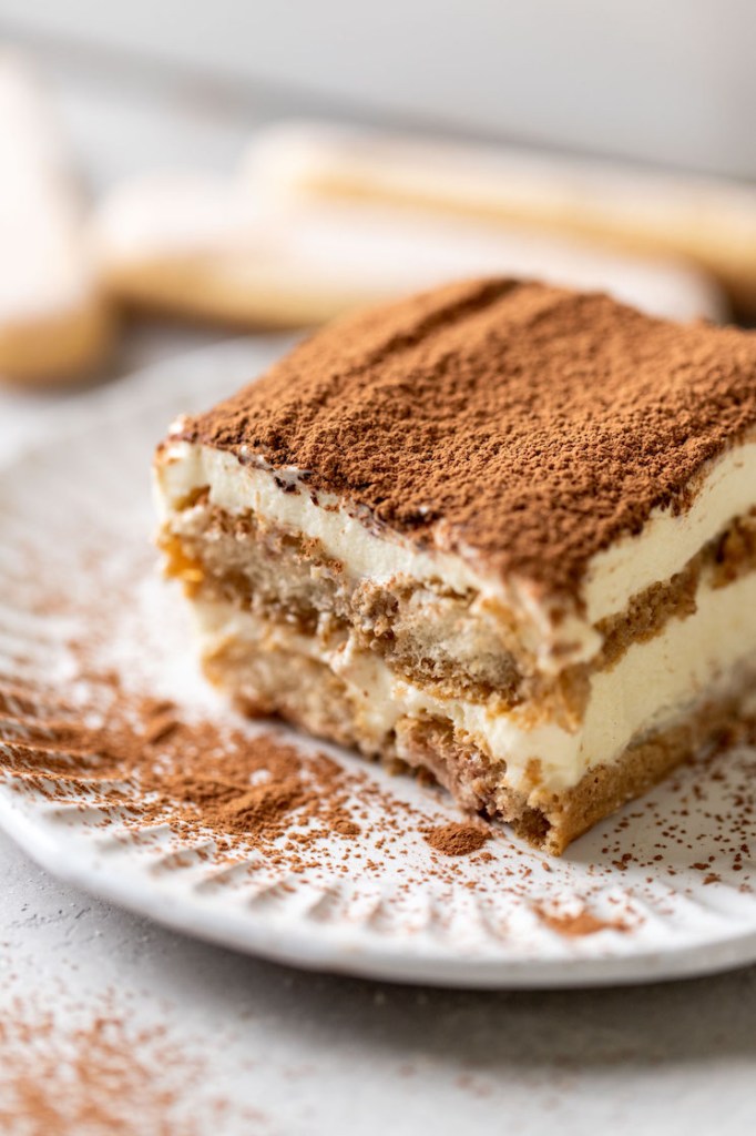 Una rodaja de tiramisú sobre un plato decorativo blanco.  Tres dedos femeninos descansan al fondo.