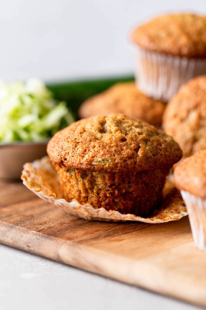 Un primer plano de un muffin de calabacín con el forro del muffin bajado.  Está sobre una tabla para cortar madera con más muffins rodeándolo. 